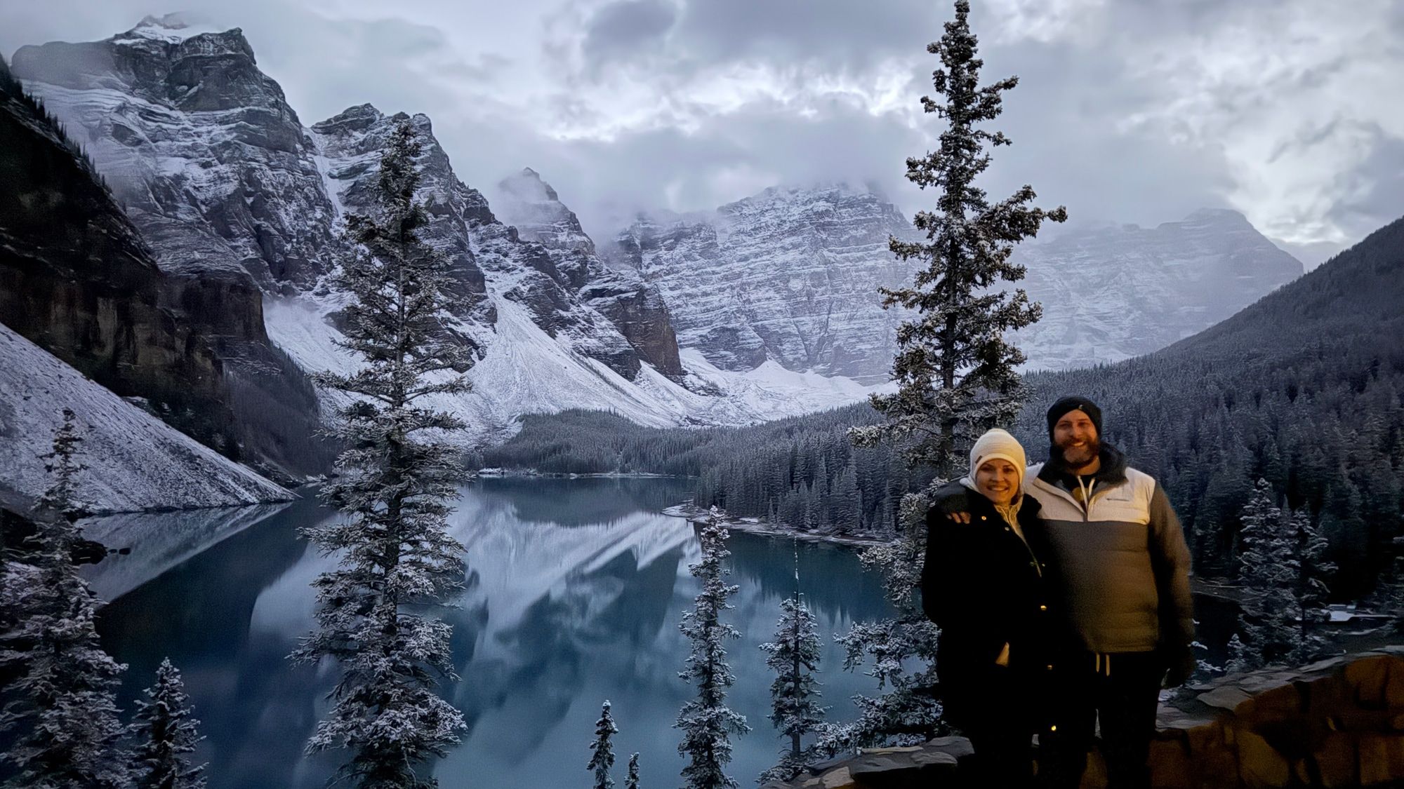 Leah and a man standing beside a lake surrounded by snowy mountains and trees